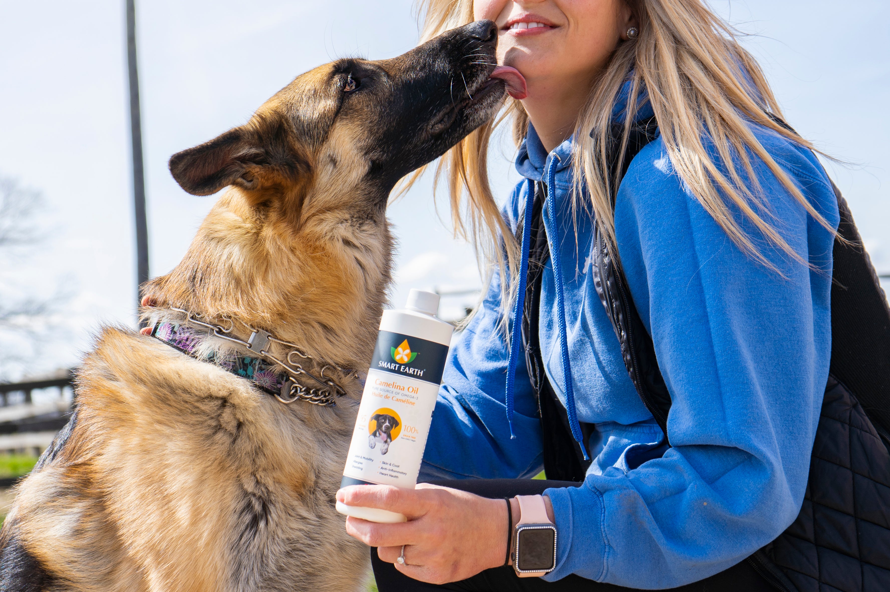 German shepherd licking its owner while she holds a bottle of Smart Earth Camelina Oil