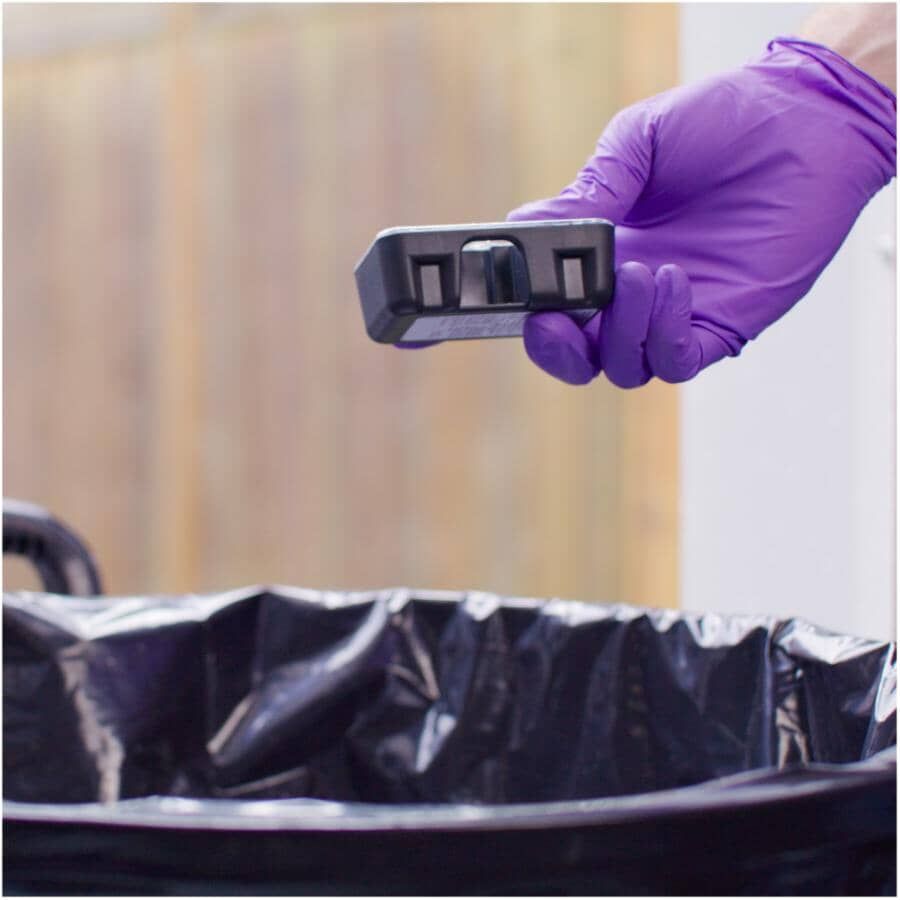 Gloved hand placing a Tomcat disposable mouse bait station beside a garbage bag